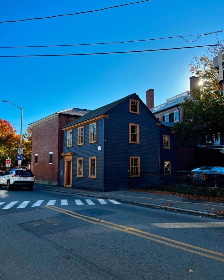 A side view of the Daniel Bray House in Salem, Massachusetts, with its dark siding and golden trim, located at a street intersection with crosswalks and parked cars under a bright blue sky.