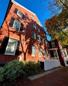 Side perspective of The Salem Inn's West House in Salem, Massachusetts, highlighting its red brick exterior, multiple windows with black shutters, and surrounding greenery.