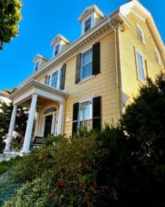 A side view of the John P. Peabody House in Salem Massachusetts, featuring its yellow exterior, white columned porch, black shutters, and well-tended greenery with red and green foliage.