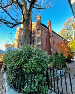 A side angle of the Salem Witch Museum in Salem, Massachusetts, showcasing its Gothic windows framed by lush greenery, vibrant fall trees, and a wrought iron fence under a clear blue sky.