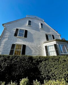 A side view of the Ropes Mansion in Salem, Massachusetts, showing its white clapboard siding, black shutters, and a large bay window above a neatly trimmed hedge under a bright blue sky.