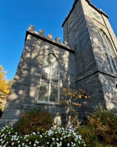 A side view of the First Church of Salem in Massachusetts, showing a Gothic-style stone tower with arched windows, surrounded by a garden of white flowers, autumn trees, and greenery under a bright blue sky.