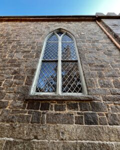 A tall, arched Gothic-style window on the stone wall of the First Church of Salem in Massachusetts, featuring diamond-patterned glass panes and a weathered stone exterior under a clear blue sky.