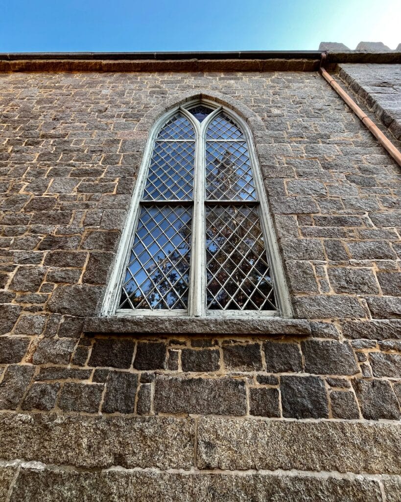 A tall, arched Gothic-style window on the stone wall of the First Church of Salem in Massachusetts, featuring diamond-patterned glass panes and a weathered stone exterior under a clear blue sky.