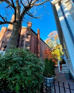 The Salem Witch Museum in Salem, Massachusetts, seen from the side, with its Gothic architecture partially obscured by a tree and lush greenery. A residential building with a staircase and pumpkins on the porch is visible in the background.