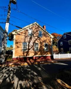 A side view of the Jonathan Whipple House in Salem, Massachusetts, highlighting its yellow siding, tall windows, and shadows cast by nearby trees under a clear blue sky.