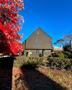 A side view of the historic John Ward House in Salem, Massachusetts, showcasing its dark wooden exterior, diamond-pane windows, and steep gables surrounded by a vibrant red-leaved tree under a bright blue sky.