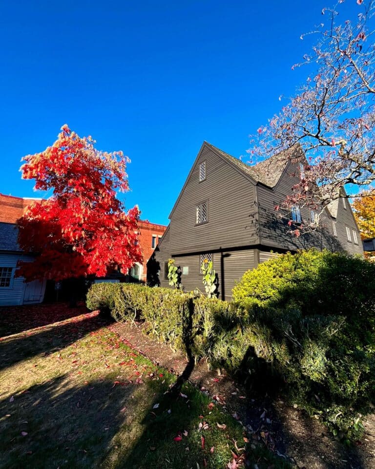 The side angle of the John Ward House in Salem, Massachusetts, showcasing its dark wooden exterior and lattice windows, with vibrant red and green foliage framing the scene under a clear blue sky.