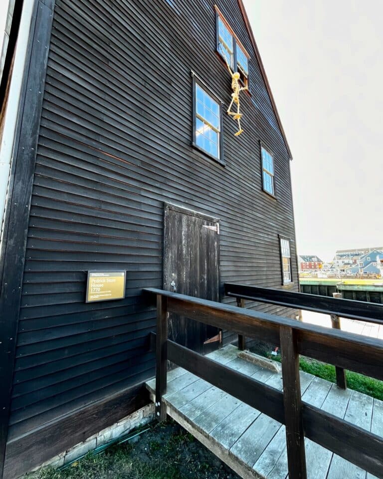 Side view of the Pedrick Store House in Salem, Massachusetts, part of the Salem Maritime National Historic Site, featuring its black siding, a wooden ramp, and a skeleton decoration.