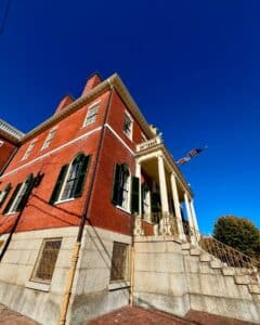 A side view of the Salem Custom House in Salem, Massachusetts, highlighting its red brick facade, tall white columns, gold-trimmed staircase, green shutters, and a waving American flag under a deep blue sky.