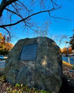 Side angle of a large granite memorial stone featuring a plaque honoring the Twenty Third Regiment, surrounded by autumn foliage and a bright blue sky in Salem, Massachusetts.
