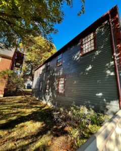 Side view of the West India Goods Store in Salem, Massachusetts, featuring dark green wooden siding, red-trimmed windows, and a tree-lined yard under a bright blue sky.