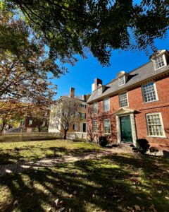 The Derby House in Salem, Massachusetts, alongside other historic homes, with its red brick Georgian architecture, green door, and a tree-lined yard under a bright blue sky.