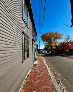 A perspective view of the Penn Townsend Manor in Salem, Massachusetts, featuring a gray exterior, a vibrant teal door, red brick sidewalk, and a tree-lined street under a bright blue sky.