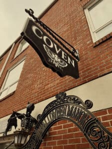 Close-up view of the Coven boutique sign featuring a moth emblem, with Halloween Manor decorations below, including skeleton statues and vintage lanterns against a red brick building in Salem, Massachusetts.