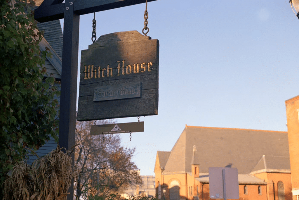 The wooden sign for the Witch House in Salem, Massachusetts, with a backdrop of trees and a historic building at sunset.