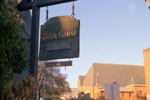 The wooden sign for the Witch House in Salem, Massachusetts, with a backdrop of trees and a historic building at sunset.