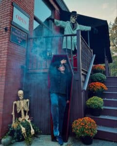 A person stepping out of a coffin with a surprised expression, while a skeleton mannequin dressed in a trench coat and hat points from above, surrounded by fall-themed decor in Salem, Massachusetts.