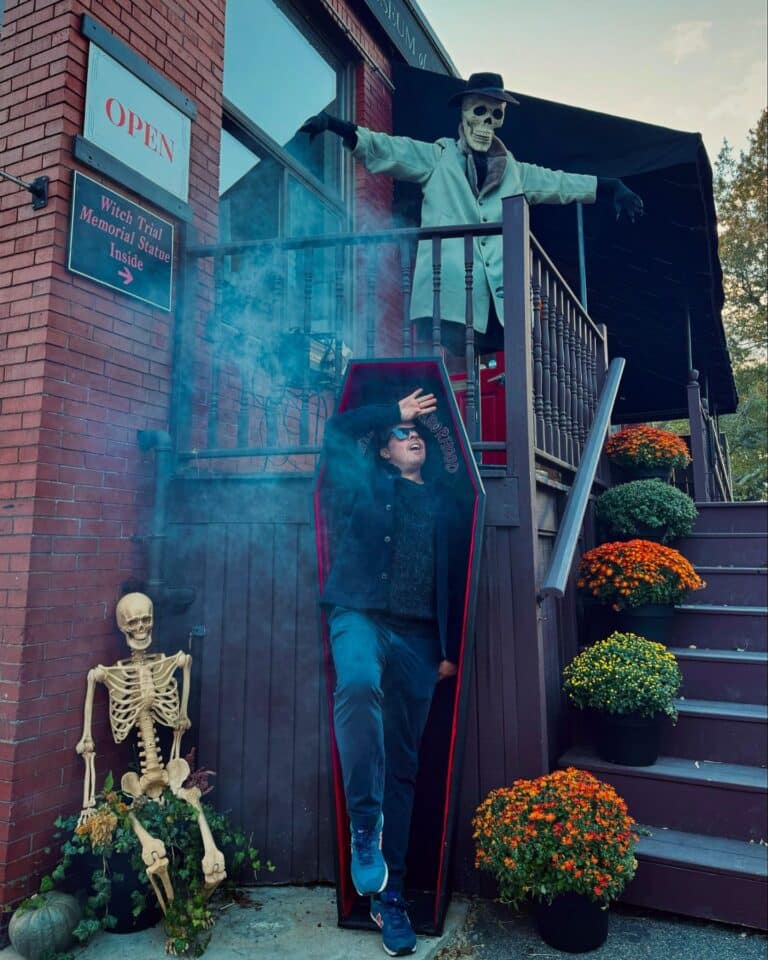 A person stepping out of a coffin with a surprised expression, while a skeleton mannequin dressed in a trench coat and hat points from above, surrounded by fall-themed decor in Salem, Massachusetts.