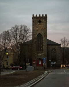 St. Peter’s-San Pedro Episcopal Church in Salem, Massachusetts, a historic stone building with Gothic windows and a square tower with battlements. The entrance has red doors, and the church sits along a quiet street at dusk.
