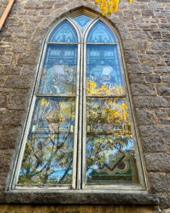 A close-up of a tall arched stained glass window on the First Church of Salem in Massachusetts, showing intricate artwork and vibrant reflections of trees with golden autumn leaves.