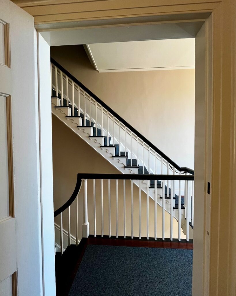 A view of a staircase in the Salem Custom House, Salem, Massachusetts, seen through a doorway, featuring white railings, dark carpet, and neutral-colored walls.