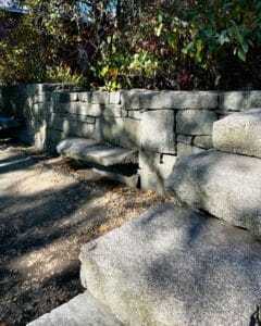 A section of the Salem Witch Trials Memorial in Salem, Massachusetts, featuring a stone wall and engraved benches set amidst leafy greenery. Sunlight and shadows create a serene and reflective atmosphere.