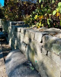 A section of the Salem Witch Trials Memorial in Salem, Massachusetts, showing a stone wall with greenery growing along it. Part of a memorial stone engraved with "Samuel" is visible, adorned with small tokens and flowers.