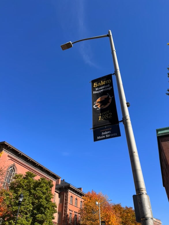 A tall streetlight with a banner reading 'Salem Haunted Happenings Since 1982' against a clear blue sky, with colorful autumn trees and red brick buildings in the background in Salem, Massachusetts.