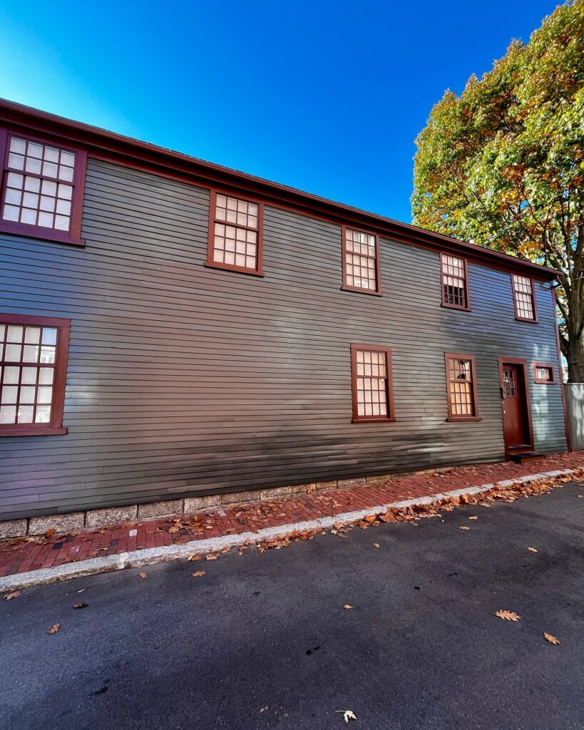 Street-facing view of the West India Goods Store in Salem, Massachusetts, showcasing its dark green wooden siding, red-trimmed windows, and brick sidewalk under a bright blue sky.