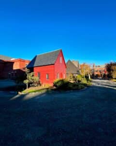The vibrant red Quaker Meeting House in Salem, Massachusetts, bathed in sunlight, surrounded by greenery and nearby historic buildings under a brilliant blue sky.