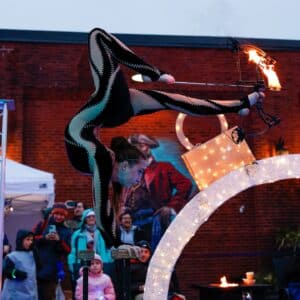 An acrobat in a sleek black costume balances on her hands while shooting a flaming arrow with her feet during an outdoor performance in Salem, Massachusetts.