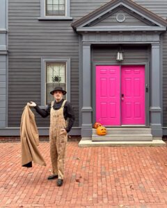 A person dressed in patterned overalls, a black shirt, and a black hat stands confidently on a brick pathway in front of a gray house with bright pink doors and pumpkins on the steps in Salem, Massachusetts.