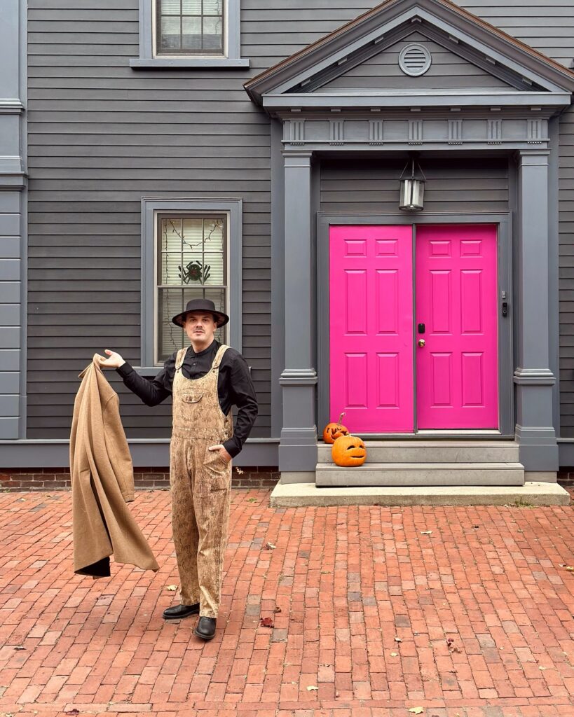A person dressed in patterned overalls, a black shirt, and a black hat stands confidently on a brick pathway in front of a gray house with bright pink doors and pumpkins on the steps in Salem, Massachusetts.
