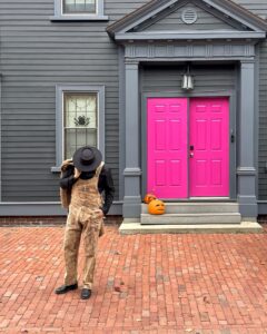 A man wearing patterned overalls, a black shirt, and a black hat poses thoughtfully while tipping his hat on a red-brick path in front of a house with vibrant pink double doors and jack-o'-lanterns on the steps in Salem, Massachusetts.