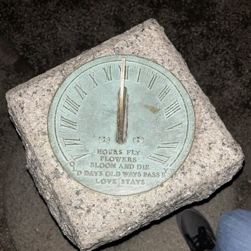 A sundial made of metal with Roman numerals sits atop a stone base at Ropes Mansion Garden. The inscription reads: "HOURS FLY, FLOWERS BLOOM AND DIE, OLD DAYS OLD WAYS PASS BY, LOVE STAYS."