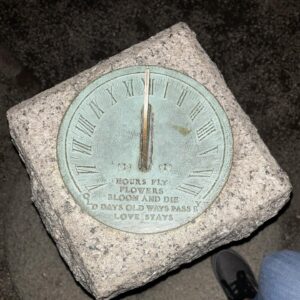 A sundial made of metal with Roman numerals sits atop a stone base at Ropes Mansion Garden. The inscription reads: "HOURS FLY, FLOWERS BLOOM AND DIE, OLD DAYS OLD WAYS PASS BY, LOVE STAYS."