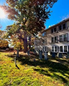 The Old Burying Point Cemetery in Salem, Massachusetts, featuring historic gravestones, a large tree with autumn foliage, and sunlight streaming near a wooden building.
