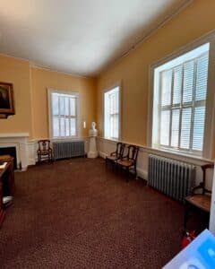 A corner of the Collector's Office in the Salem Custom House, Salem, Massachusetts, featuring large windows with shutters, a bust on a pedestal, wooden chairs, and a fireplace.