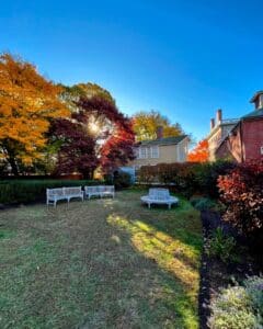A peaceful garden behind the Andrew-Safford House in Salem Massachusetts, featuring white benches, vibrant autumn trees, and golden sunlight streaming through the foliage.