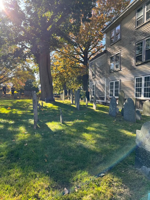 A grassy area at the Old Burying Point Cemetery with gravestones scattered among the tall trees, casting long shadows on a sunny day in Salem