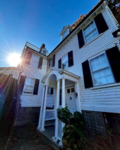 The side entrance of the Ropes Mansion in Salem, Massachusetts, featuring a white arched portico with columns, black shutters, and the sun shining brightly above the historic colonial house.