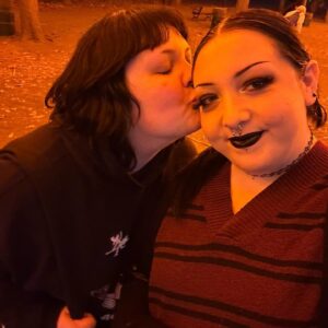 A close-up photo of two people at Salem Common. One person is giving a gentle kiss on the cheek of the other, who is smiling at the camera. The lighting has an orange hue, creating a warm and cozy atmosphere as leaves are scattered on the ground in the background.