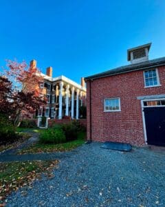 View of the Andrew-Safford House in Salem Massachusetts with its grand columns, flanked by a smaller red brick building under a clear blue sky.