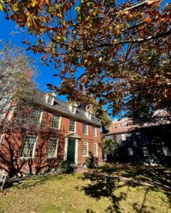 The Derby House in Salem, Massachusetts, featuring red brick Georgian architecture, a green door, and autumn leaves, with a neighboring historic home visible under a bright blue sky.