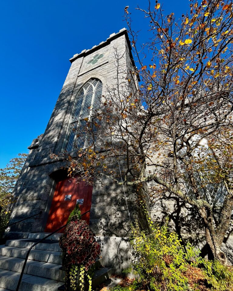 The First Church of Salem in Massachusetts, a historic stone building with red wooden doors, tall arched windows, and a castle-like tower, surrounded by trees with autumn leaves under a bright blue sky.