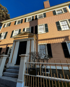 The Hawkes House in Salem, Massachusetts, showcasing its Federal-style architecture with peach clapboard siding, white trim, black shutters, and a decorative entrance framed by columns under a bright blue sky.