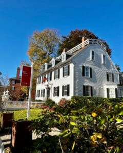 The Ropes Mansion in Salem, Massachusetts, a white colonial house with black shutters and a red Peabody Essex Museum sign in front, surrounded by lush greenery, colorful gardens, and fall trees under a bright blue sky.