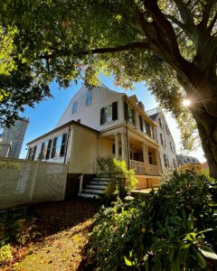 The Ropes Mansion in Salem, Massachusetts, with sunlight filtering through tree branches, casting shadows on the house and surrounding plants.