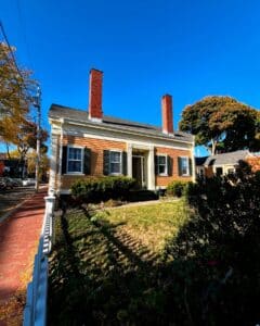 The Jonathan Whipple House in Salem, Massachusetts, showcasing a classic mid-19th century design with a yellow-painted exterior, green shutters, and two prominent brick chimneys against a bright blue sky.